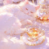 A fluffy white kitten rests on a pink surface next to a glass cup filled with golden liquid.