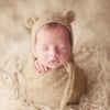 A newborn baby sleeping peacefully wearing a knitted bear hat and wrap set on a light, fluffy surface.