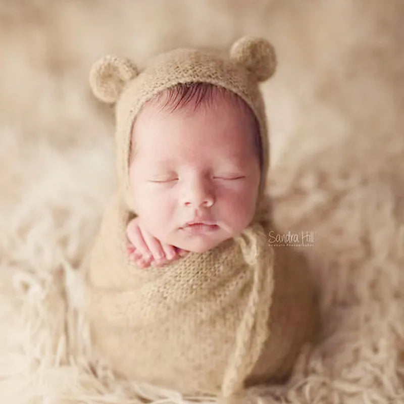 A newborn baby sleeping peacefully wearing a knitted bear hat and wrap set on a light, fluffy surface.