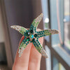 A sparkling green and gold rhinestone turtle brooch pin displayed on a hand.