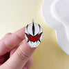 A hand holds a silver enamel pin featuring a red and black dinosaur ranger helmet design against a light background.
