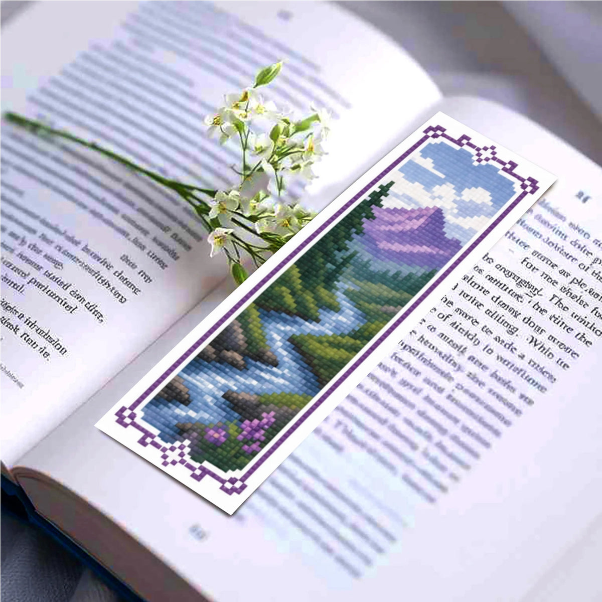 A DIY cross-stitch bookmark craft kit featuring a floral design and purple and blue patterned paper inside an open book.
