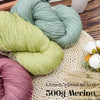 A pile of light green and pink merino wool yarn skeins resting on a woven fabric background.