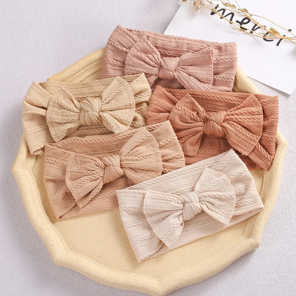 A set of three beige and pink soft stretch baby headbands with bows displayed on a light yellow tray.
