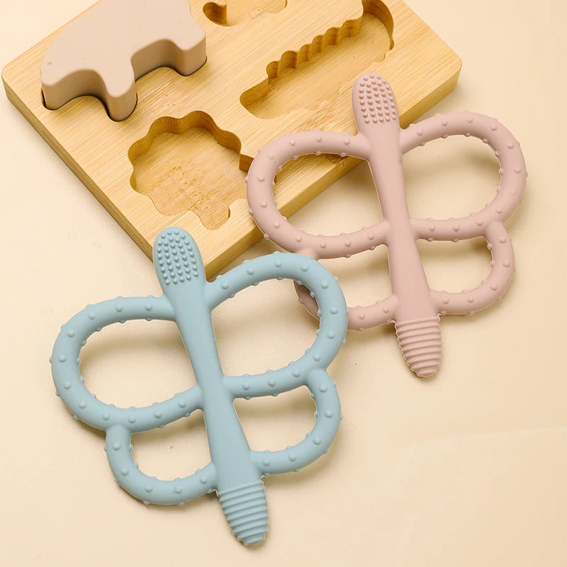 A set of soft silicone butterfly teethers in pink and light blue, resting on a wooden tray.