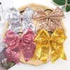 A collection of five glitter sequin pigtail bows in pink, silver, bronze, and gold are displayed on a wooden tray.