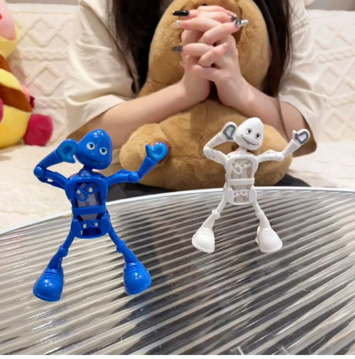 A blue wind-up dancing robot toy is displayed on a glass surface next to a white clockwork robot.