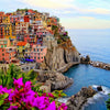 A colorful cliffside village overlooking the sea with a small cove and vibrant purple flowers in the foreground.