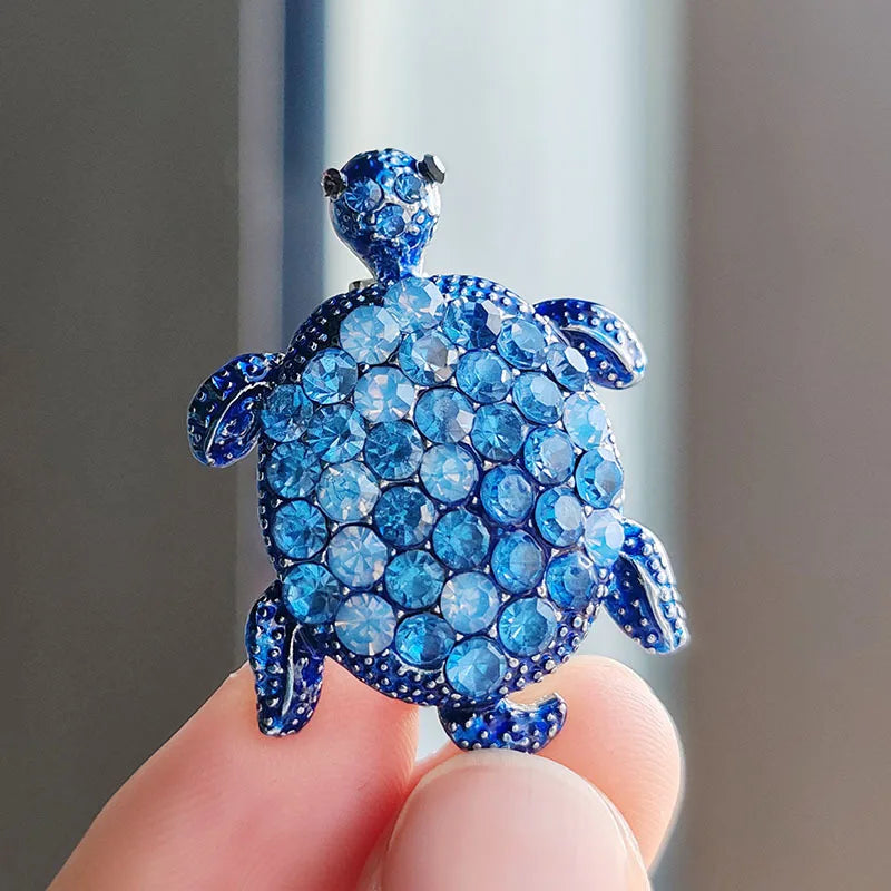 A detailed blue rhinestone turtle brooch pin held by a person's fingers.