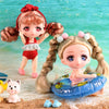 A small doll with pink hair and red bow is shown next to a blue inflatable ring on a sandy surface.
