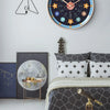 A round wall clock with a dark background and colorful markings, displayed above a modern bed.