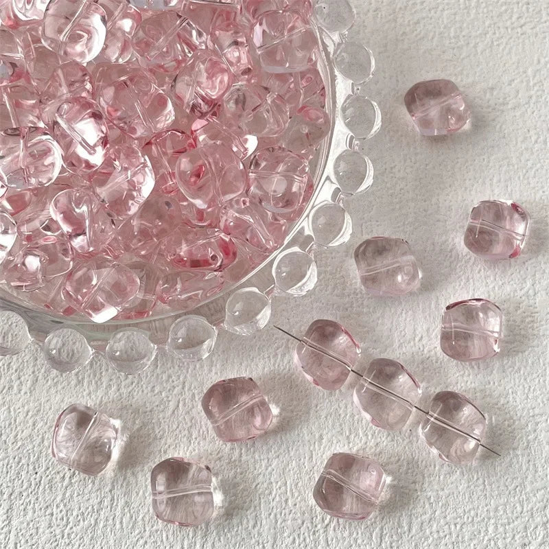 A clear glass bowl filled with pink irregular square beads for jewelry making scattered on a white surface.