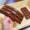 A hand holds a brown, realistic chocolate squishy toy with a grid pattern on a woven tray.