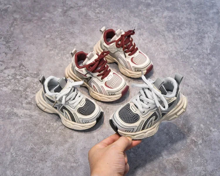 A hand holds a pair of red and white breathable mesh kids running sneakers with cushioned soles.