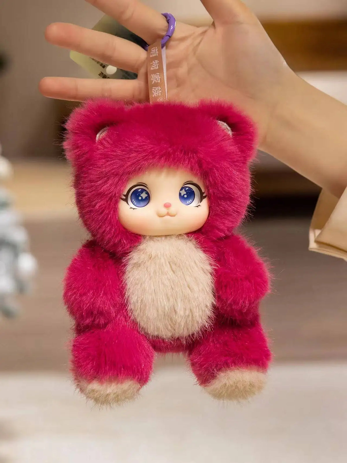 A pink plush pendant featuring a rabbit face with blue eyes and a fluffy texture, held by a hand.