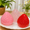 A bright pink and red strawberry squishy toy resting on a white plate with green plants in the background.