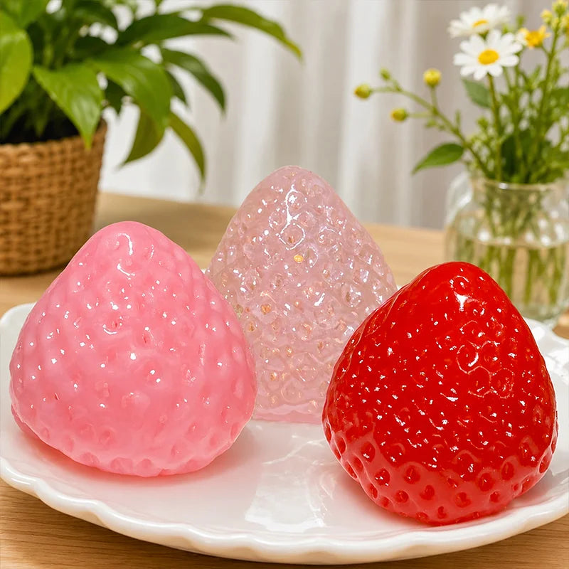 A bright pink and red strawberry squishy toy resting on a white plate with green plants in the background.