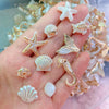 A hand displays various iridescent and pearlescent ocean animal acrylic beads including starfish, shells, and a fish.