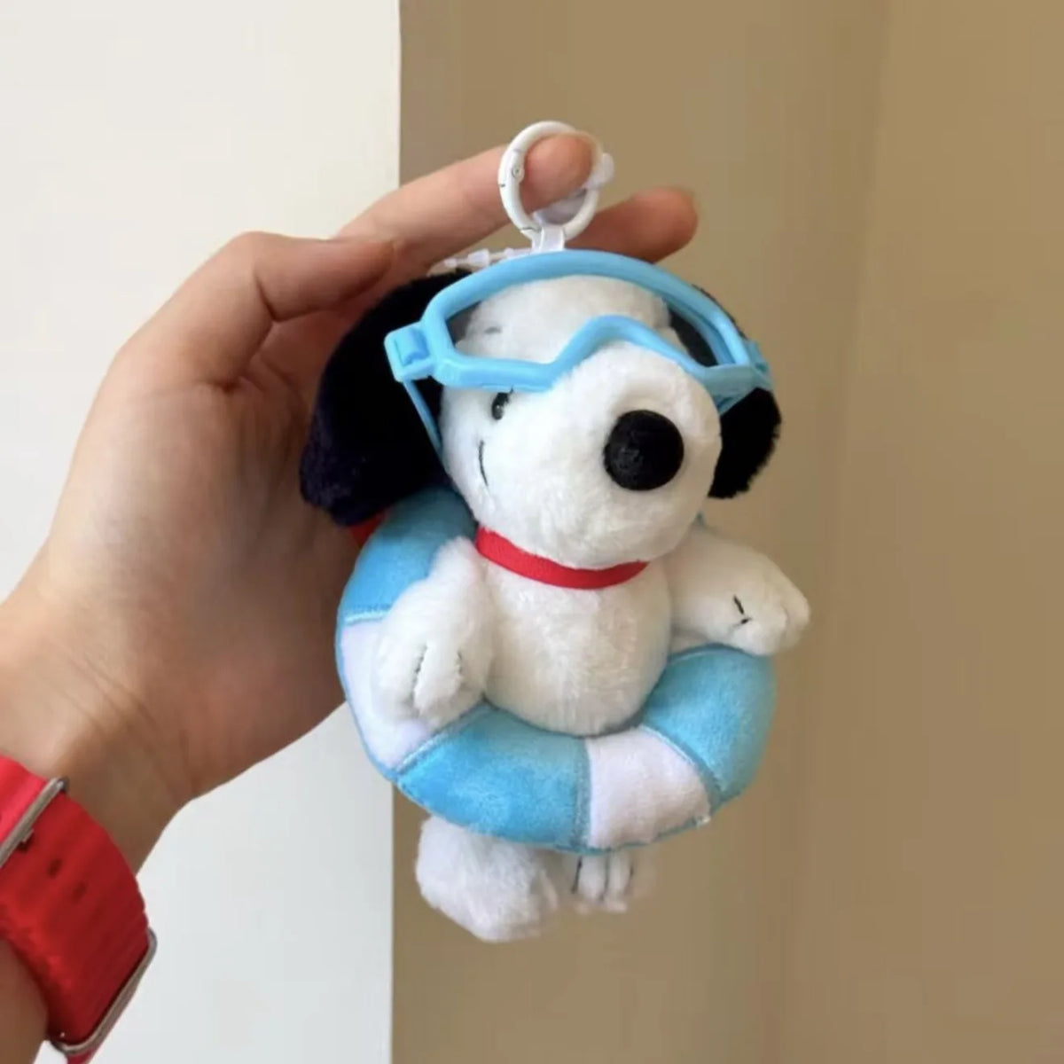 A white Snoopy plush charm wearing blue goggles and a red collar being held by a hand.