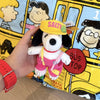 A hand holds a Snoopy plush keychain charm with a yellow hat and pink outfit against a colorful background.