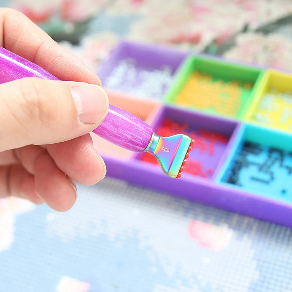 A hand holds a purple diamond painting pen with a metal tip over a colorful plastic palette containing resin diamonds.