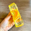 A hand holds a clear yellow ice cube shaped like a slice of orange on a wooden surface.