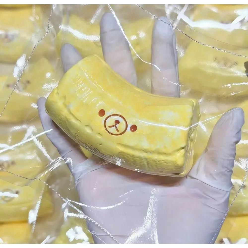 A hand holds a yellow banana bear squishy stress relief toy with a butter-textured silicone finish.
