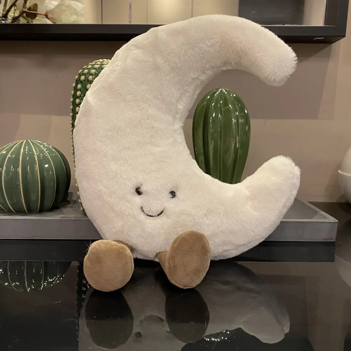 A white crescent moon plushie with a smiling face is displayed next to two green cactus-shaped objects.