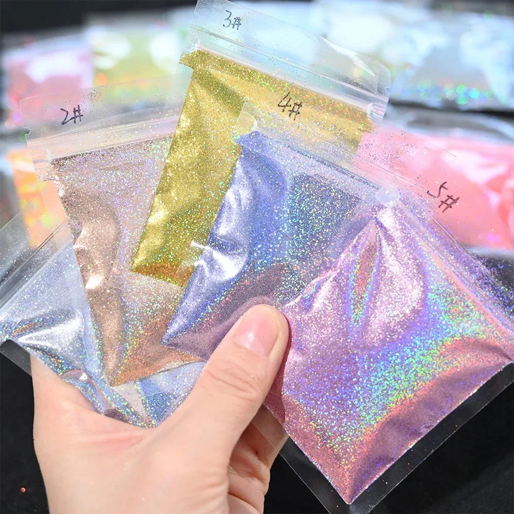 A hand holds several small plastic bags of holographic rainbow nail glitter in various shades.