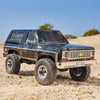 A black 1:24 scale Chevrolet K5 Blazer RC crawler parked on sandy terrain under a clear blue sky.
