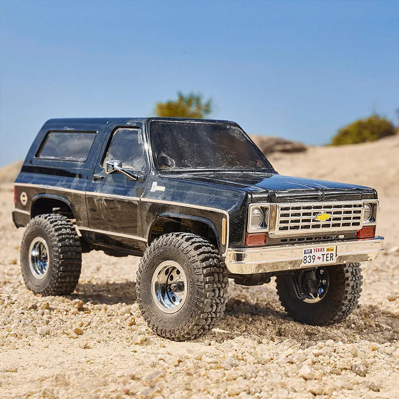 A black 1:24 scale Chevrolet K5 Blazer RC crawler parked on sandy terrain under a clear blue sky.