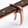 A close-up view of a bronze metal guitar capo with a pick holder attached, resting on a wooden fretboard.