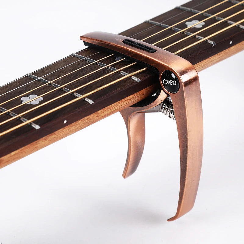 A close-up view of a bronze metal guitar capo with a pick holder attached, resting on a wooden fretboard.