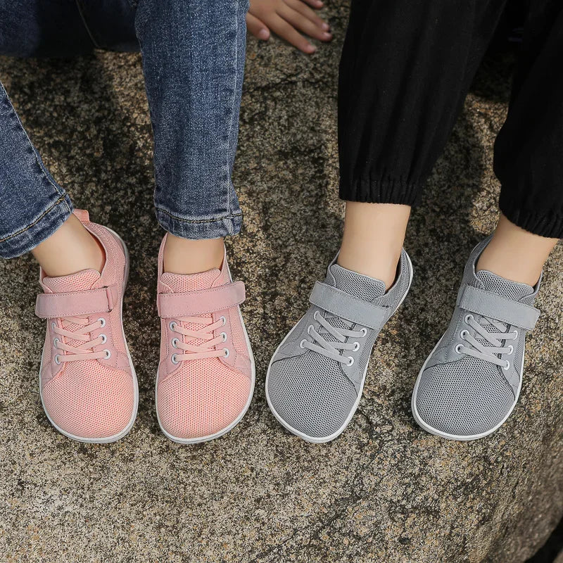 A pair of pink and gray minimalist barefoot sneakers worn by a child against a textured background.