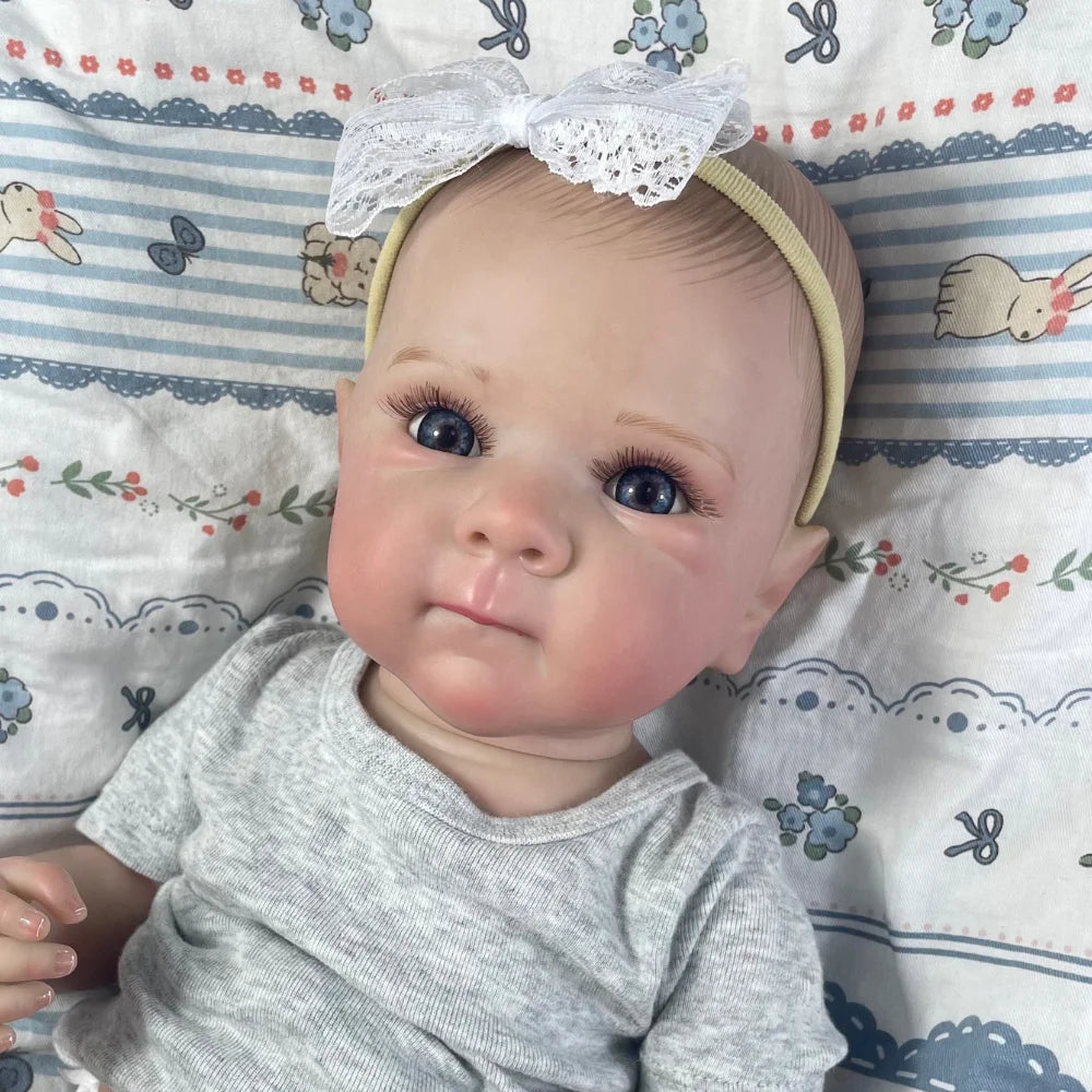 A realistic reborn baby doll with painted hair wearing a gray shirt, lying on a patterned fabric.
