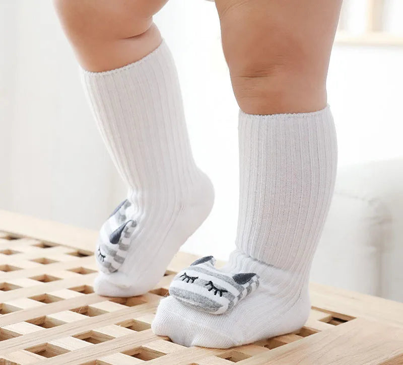 A pair of white cotton socks with cartoon cat designs worn on a baby's foot.