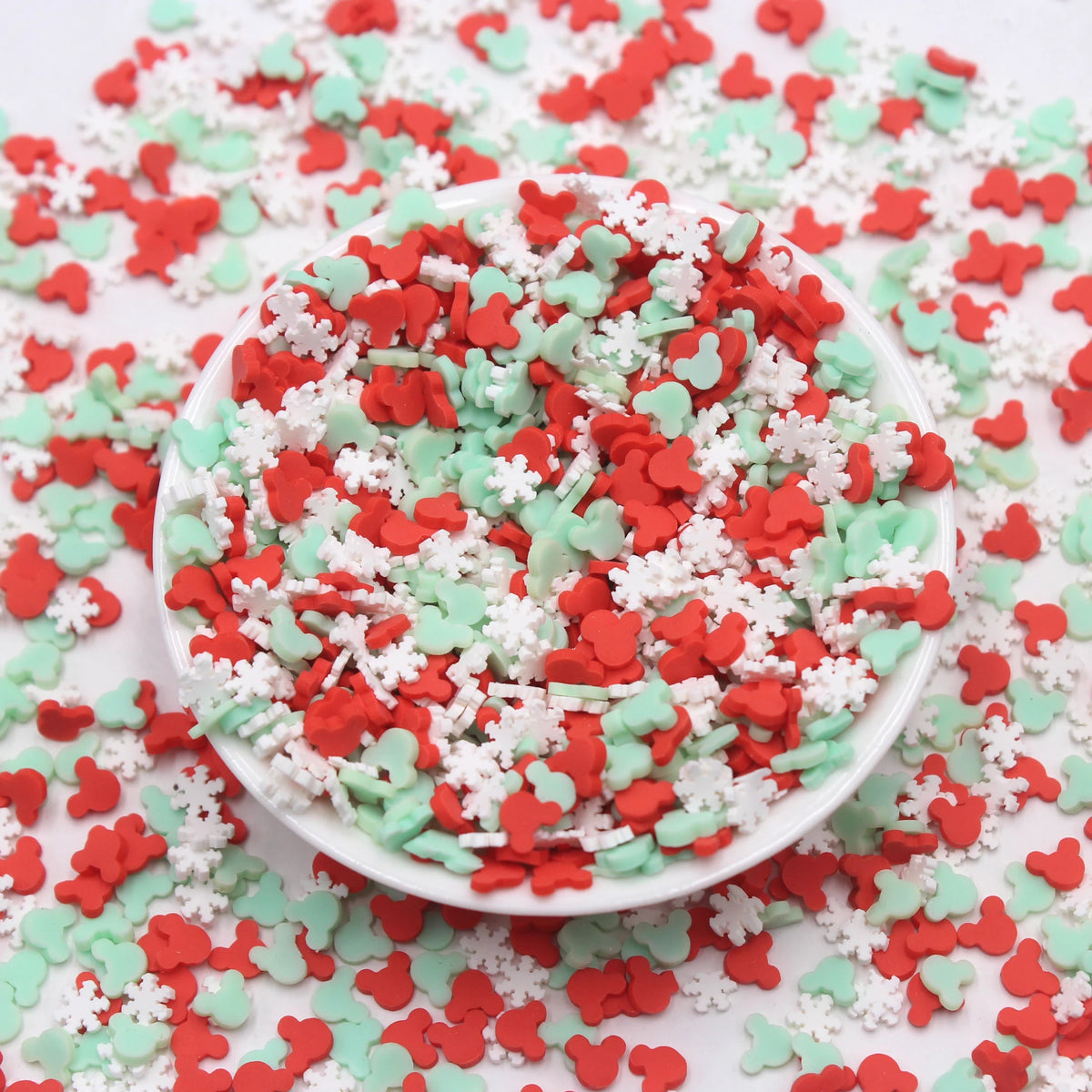 A white bowl filled with red and green cartoon polymer clay slices for slime and craft projects scattered on a white surface.