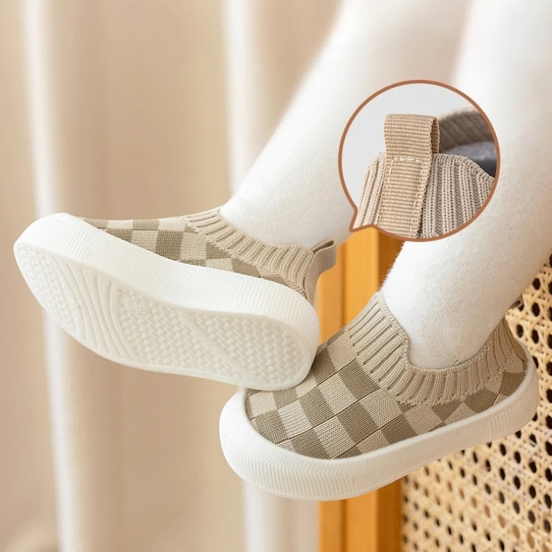 A pair of beige and white plaid slip-on toddler shoes for first walkers shown on a person's foot.