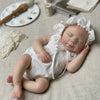 A newborn baby doll made of silicone with a white lace bonnet and outfit lying on a beige cloth.