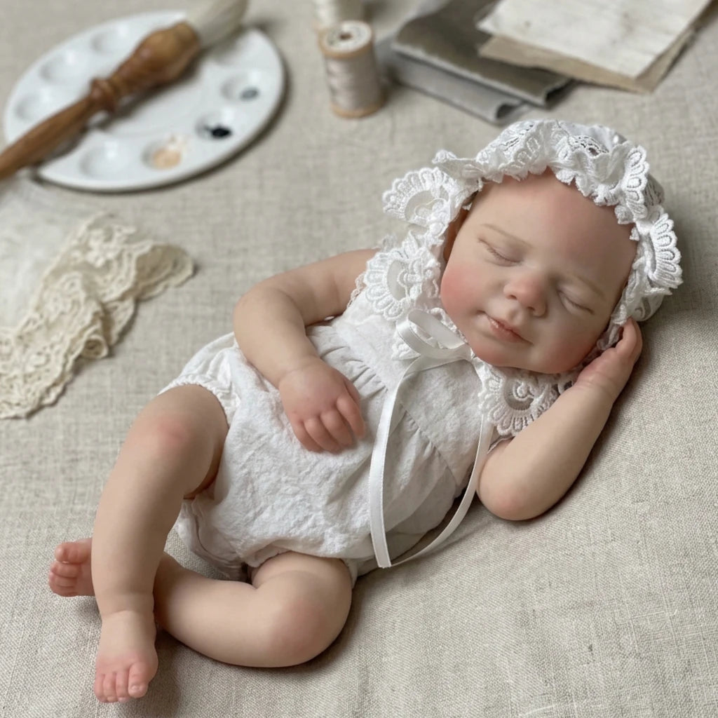 A newborn baby doll made of silicone with a white lace bonnet and outfit lying on a beige cloth.