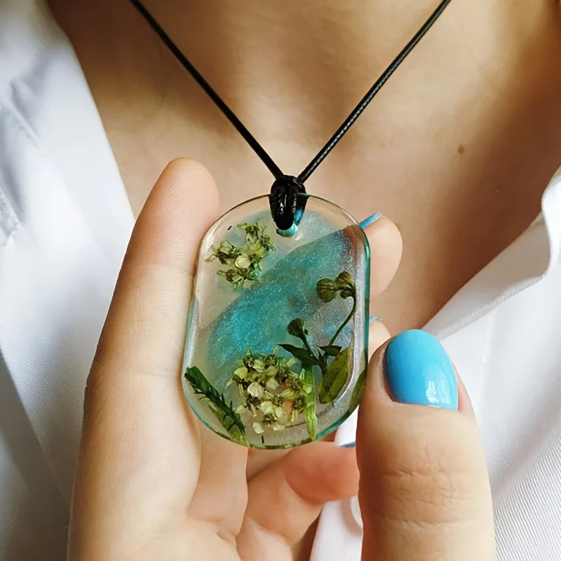 A hand holds a clear epoxy resin mold featuring green and white floral elements, suspended from a black cord.