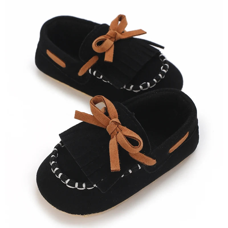 A pair of black baby first walker sneakers with brown ribbon ties and white stitching.