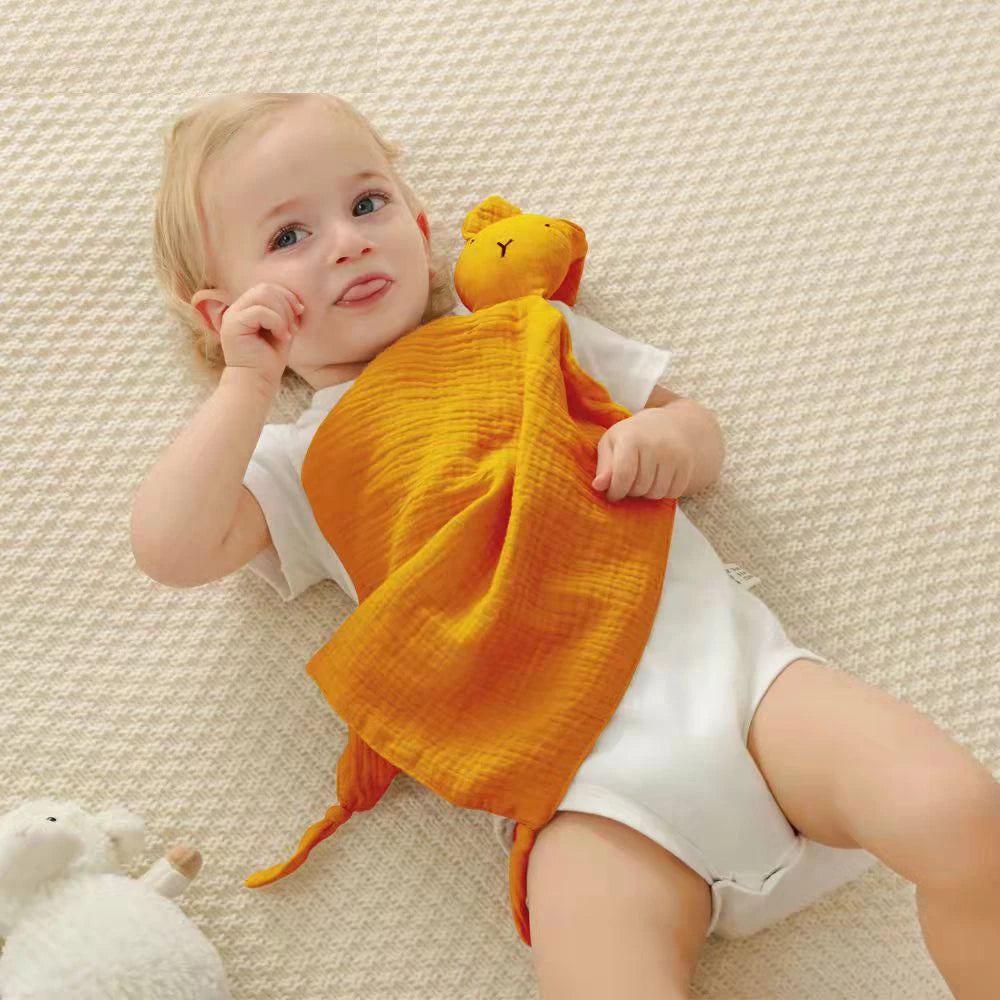 A baby is holding an orange soft cotton muslin blanket while lying on a light textured surface.