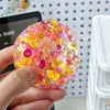 A hand holds a translucent, colorful fidget ball made of soft silicone, featuring pink, yellow, and purple hues.