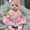 A realistic pink reborn baby doll with painted hair wearing a pink dress and white socks, sitting on a gray fur rug.