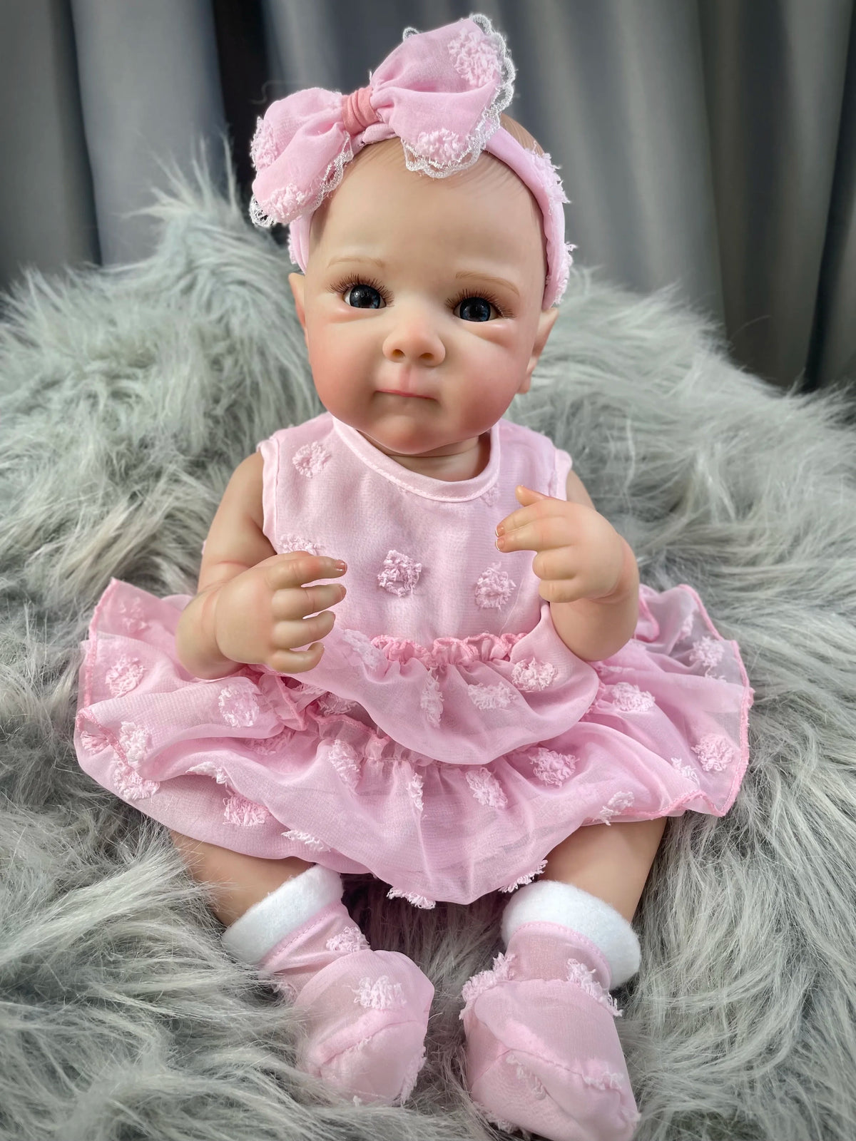 A realistic pink reborn baby doll with painted hair wearing a pink dress and white socks, sitting on a gray fur rug.