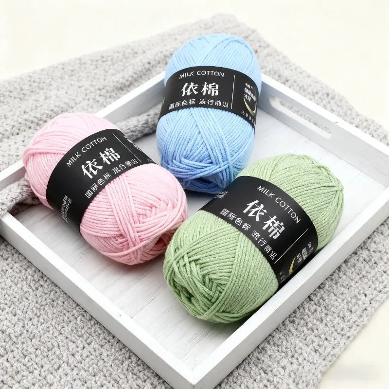 Three skeins of soft milk cotton yarn in light blue, pink, and green are displayed in a white wooden frame.