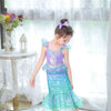 A young girl wearing a purple and teal mermaid fantasy dress with a ruffled fishtail skirt stands in front of a sheer window.