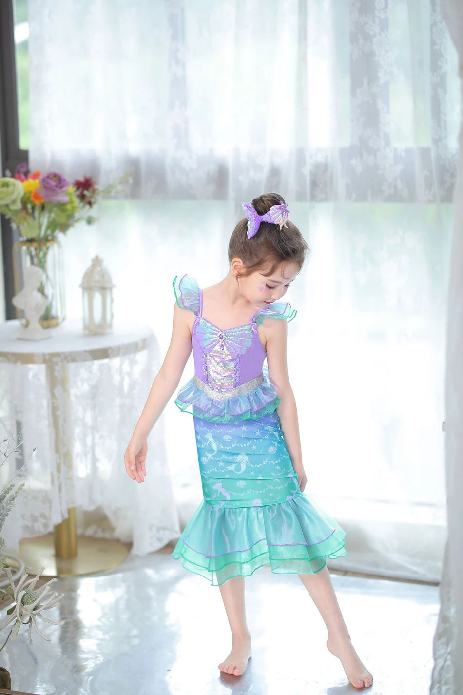 A young girl wearing a purple and teal mermaid fantasy dress with a ruffled fishtail skirt stands in front of a sheer window.