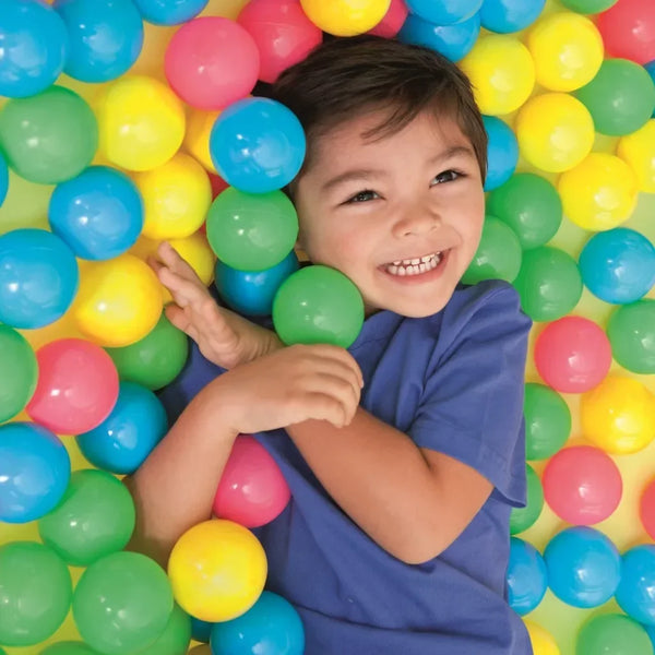 A smiling child holds a purple shirt surrounded by a bulk set of colorful plastic ocean ball pit toys.